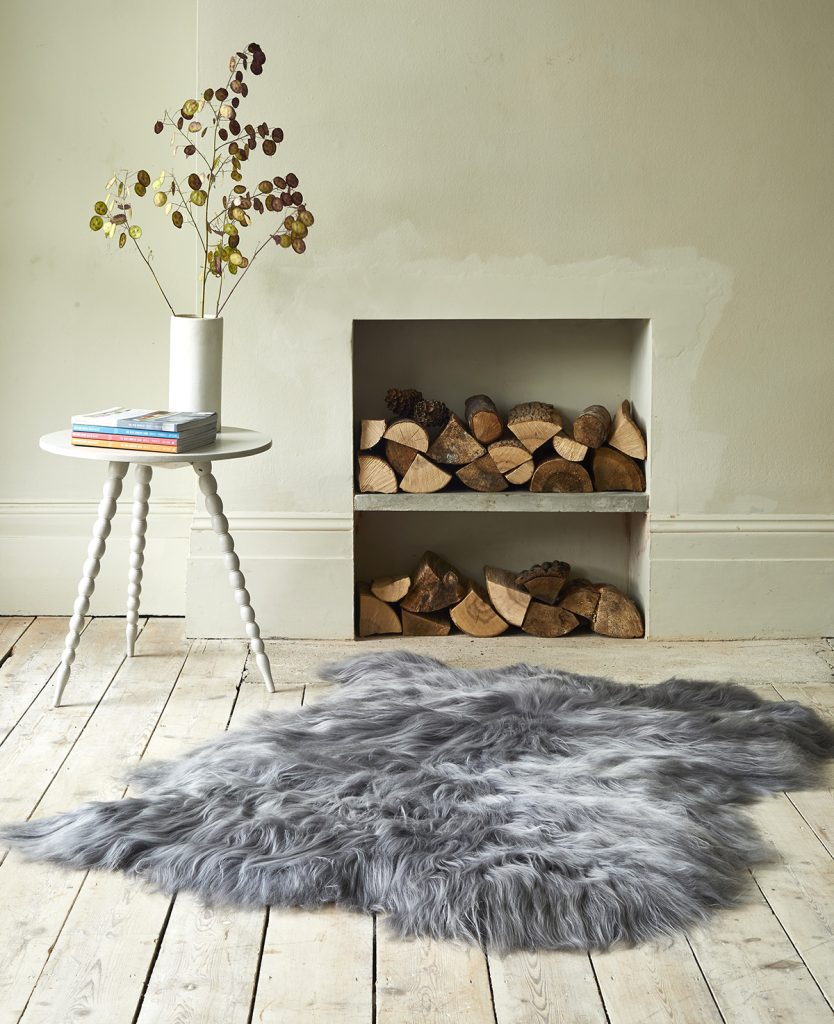 Grey sheepskin laid on a wooden floor, infront of a log store. Small table with flowers next to it.